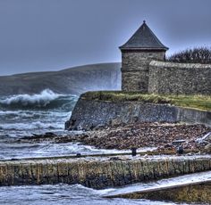 an old stone wall next to the ocean with waves crashing on it and a lighthouse in the distance