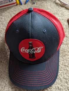 a black and red coca cola hat sitting on top of a carpet next to crayons