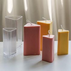 four different colored candles sitting next to each other on a white counter top in front of a curtain