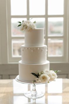 a three tiered white wedding cake with flowers on the top and bottom, in front of a window