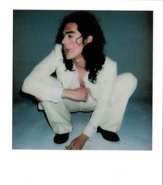 a man with long hair and glasses sitting on the ground wearing a white suit while posing for a photo