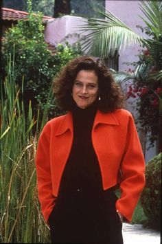 a woman in an orange coat is standing by some plants and trees with her hands on her hips