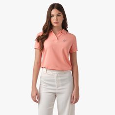 Front view of a woman wearing a peach-colored polo shirt with an embroidered logo on the chest, paired with white pants. Casual Polo Collar Tops For Work, Casual Tops With Polo Collar For Work, Classic Cotton Tops, Summer Collared Polo Shirt, Summer Casual Polo Shirt For Workwear, Casual Summer Polo Shirt For Work, Casual Relaxed Fit Pima Cotton T-shirt, Relaxed Fit Pima Cotton T-shirt With Short Sleeves, Summer Cotton Moisture-wicking Polo Shirt