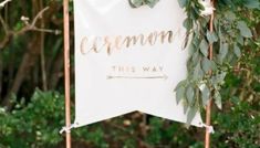 a white sign with greenery on it that says ceremony this way and is hanging in front of some trees