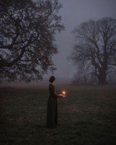 a woman holding a lit candle in a field at night with fog and trees behind her