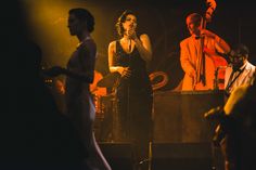 a woman in a black dress sings into a microphone while other people stand behind her