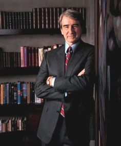 a man wearing a suit and tie standing in front of a bookshelf with his arms crossed