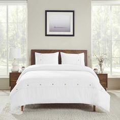 a bed with white linens and pillows in a large room next to two windows