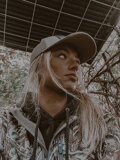 a woman with long hair wearing a hat and looking off into the distance while standing in front of some branches