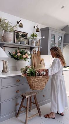 a woman standing in a kitchen next to a basket filled with flowers and cutting boards