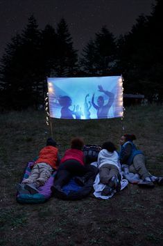 group of people sitting on the ground watching a movie at night with projection screen in background