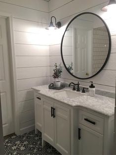 a white bathroom with black and white tile flooring, round mirror above the sink