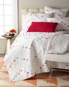 a white bed with red and blue pillows on it's headboard next to a window