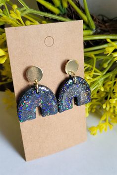 a pair of earrings with blue and purple glitters on them sitting next to yellow flowers