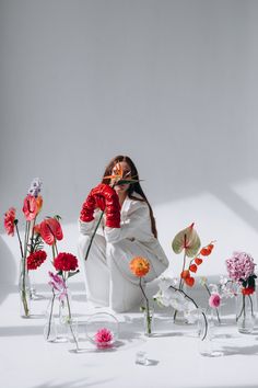 a woman sitting on the ground surrounded by vases filled with flowers and holding a flower in her hand