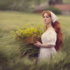 a woman with long red hair holding a basket full of yellow flowers in a field