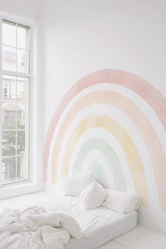 a white bed sitting under a window next to a rainbow wall mural on the wall