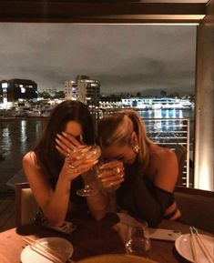 two women sitting at a table with wine glasses in front of their faces and the city lights behind them