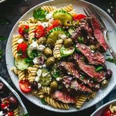 a white plate topped with pasta salad next to two bowls filled with olives, tomatoes and cucumbers