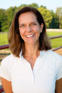 a woman in a white shirt smiling at the camera with trees and grass behind her