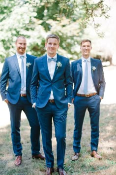 three men in blue suits standing next to each other on the grass with trees in the background