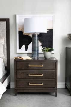 a nightstand with a lamp on top of it next to a painting and a bed