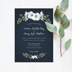 a wedding card with white flowers and greenery on the front, next to a green leafy branch