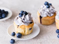 three blueberry cupcakes with frosting and fresh berries on the plate next to them