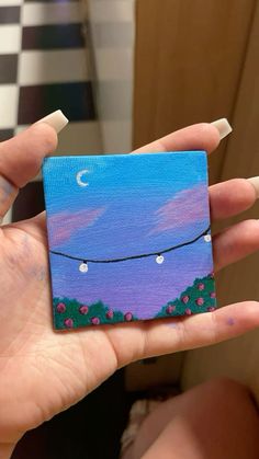 a hand holding a small square painting with string lights and stars on the night sky