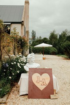 a heart shaped sign sitting on top of a gravel road next to flowers and trees