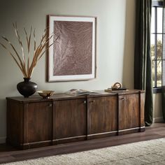 a room with a large painting on the wall next to a wooden cabinet and vase