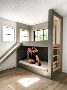 a woman sitting on top of a bunk bed in a room with stairs and windows