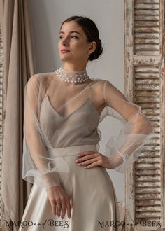 a woman wearing a dress with sheer sleeves and pearls on the neck, standing in front of a window