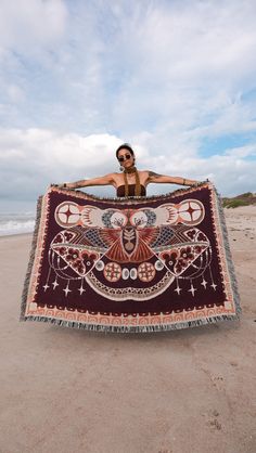 a woman sitting on top of a sandy beach holding up a large blanket with an owl design