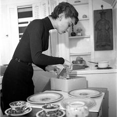 an old photo of a woman in the kitchen with plates and bowls on the table