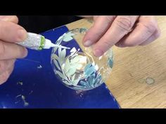 an older person painting a glass ornament with white flowers and leaves on it