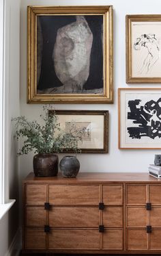 several framed pictures hang on the wall next to a dresser with vases and books