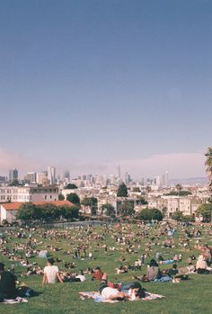 many people are laying on the grass in a park with buildings in the background and blue sky