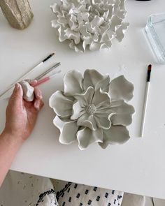 a person holding a pencil in their hand next to some flowers on a white table