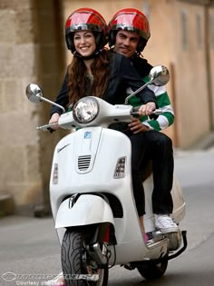 a man and woman riding on the back of a white scooter down a street