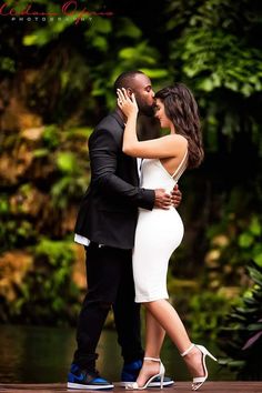 a man and woman standing next to each other in front of some water with trees behind them