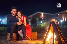 a man and woman sitting next to a campfire at night with their arms around each other