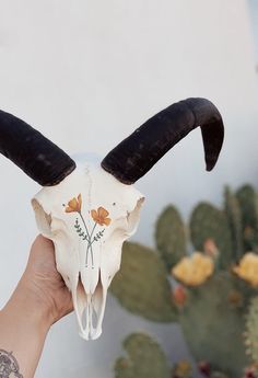 a hand holding a skull with long horns and flowers on it's face in front of a cactus