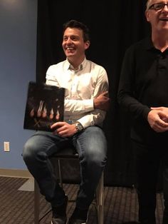 two men are sitting in chairs and one is holding a poster with the same image on it