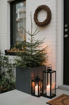 two lit candles sit next to a small christmas tree on the front porch with a wreath hanging above it