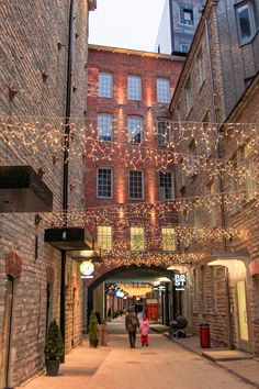 people walking down an alley way with christmas lights strung across the walkway and buildings in the background