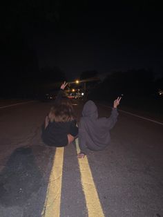 two people sitting on the side of a road at night with their hands in the air