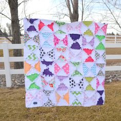 a colorful quilt hanging on a fence in front of a white fence and some trees