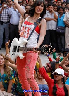 a woman in red pants and white shirt holding a guitar while standing next to a group of people