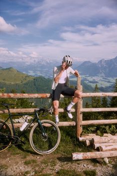 a man sitting on top of a wooden fence next to a bike and talking on a cell phone